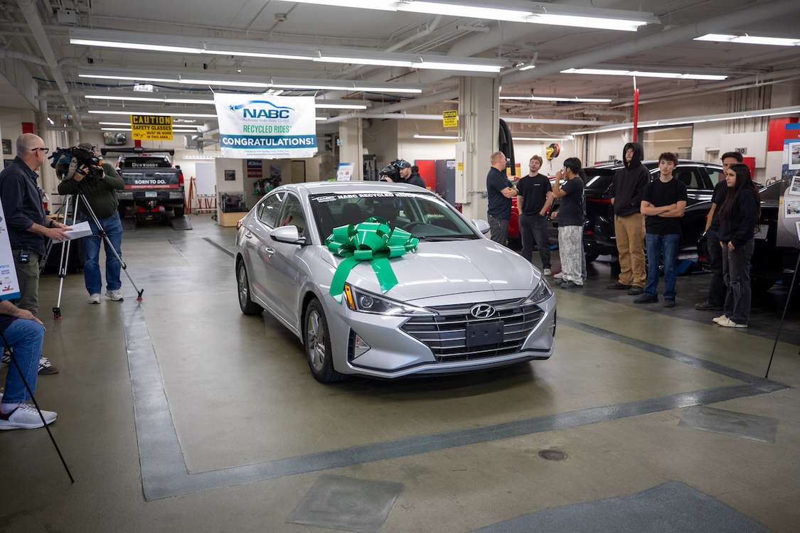 A silver Hyundai sedan adorned with a large green bow is showcased in a garage, surrounded by people and media, with a banner celebrating “NABC Recycled Rides.”